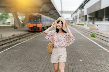 young asian woman traveler with weaving basket happy smiling and looking to a camera beside train coming background. Journey trip lifestyle, world travel explorer or Asia summer tourism concept.