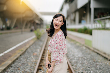 Fototapeta premium young asian woman traveler with weaving basket happy smiling looking to a camera beside train railway. Journey trip lifestyle, world travel explorer or Asia summer tourism concept.