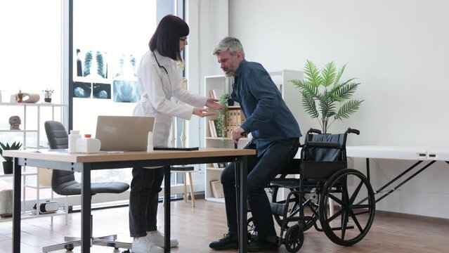 Young woman in lab coat helping mature man with walking cane to stand up from wheelchair in consulting room. Efficient therapist assisting patient in finding balance during recovery from injury.