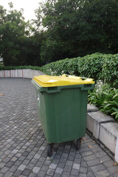 Green and yellow trash can in the public park, with flower and leaves around it.