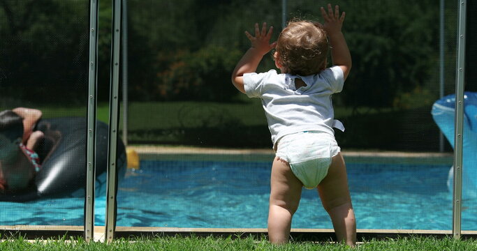 Baby child holding into swimming pool fence watching children play at pool