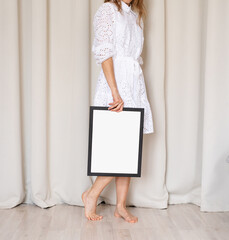 woman in dress holding blank frame, beige curtain background