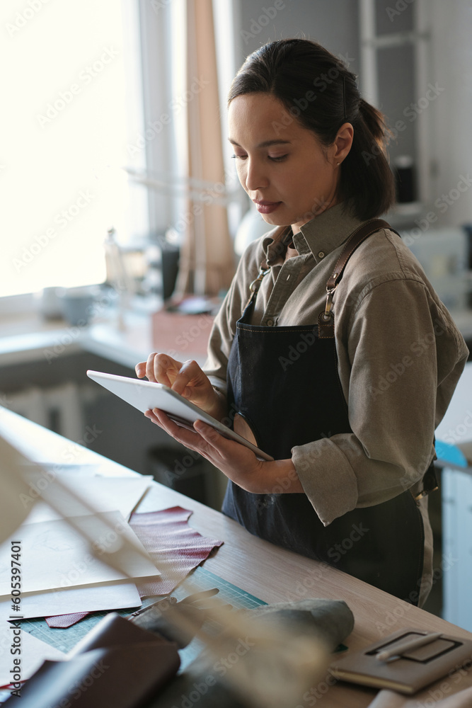 Poster young serious female artisan in workwear scrolling in tablet while standing by workplace in studio a