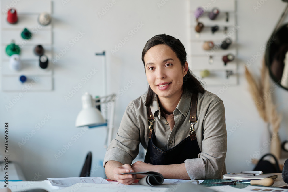 Aufkleber young smiling brunette woman in workwear sitting by table and looking at camera while working with t