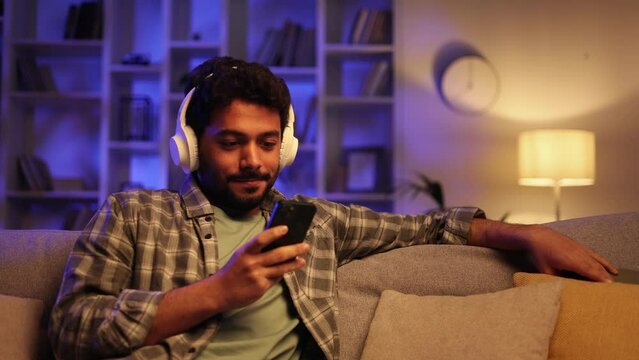 Happy young man with white headphones chilling and listening his favorite song. Arabian brunette man seat on sofa and relax after work, uses smartphone for play music, clicking on the screen.
