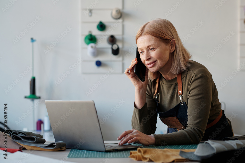 Aufkleber Smiling blond craftswoman talking on smartphone to client while looking at laptop screen and networking or discussing details of online orders