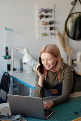 Mature smiling female tailor or leatherworker specifying details of online order while looking at laptop screen and talking on smartphone - obrazy, fototapety, plakaty