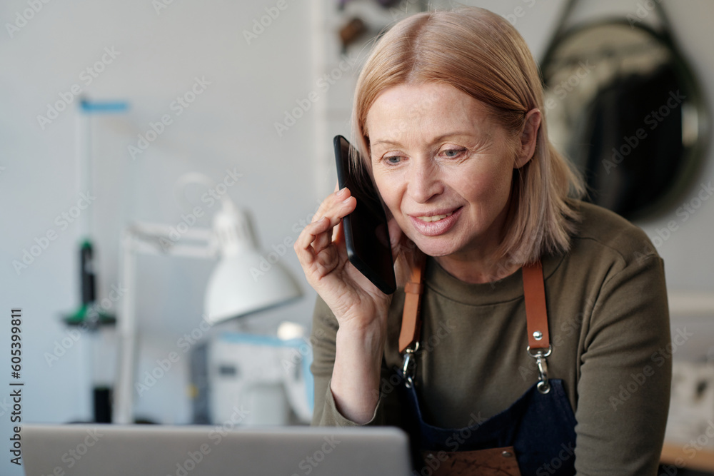 Poster smiling blond mature woman with smartphone by ear looking at laptop screen and talking to one of cli