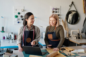 Mature blond female tailor with smartphone in hand looking at laptop screen and listening to young...