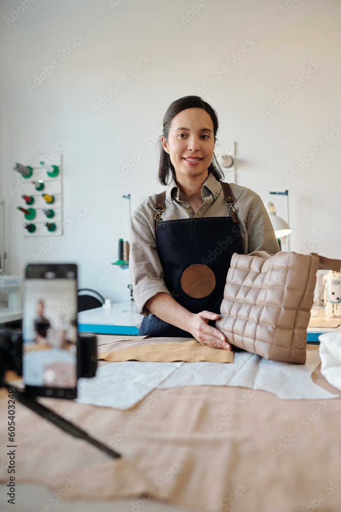 Aufkleber young smiling creative woman in apron with new handmade leather bag looking at smartphone camera whi