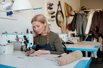 Mature craftswoman in workwear sewing new leather attire or other trendy items while creating seasonal fashion collection for her clients