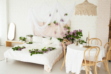 beautiful bright room in boho style with a wooden table and chairs and a large bed covered with a white bedspread and decorated with lilac flowers