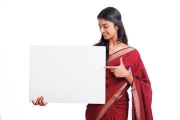 Cheerful Indian woman in traditional saree holding and pointing sign board isolated on white....