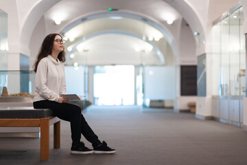 Young Caucasian woman student sitting on bench in museum visiting educational excursion. Light hall in background. Concept of cultural exhibition