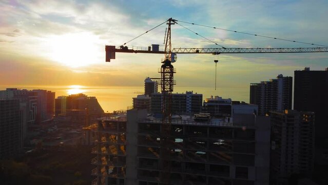 New modern apartment buildings, high-rise building with construction crane against backdrop of sunset, sun on ocean or sea. Drone view of crane construction site over city at sunset. Contractor.