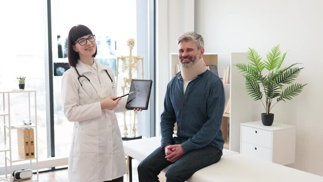 Cheerful young woman with digital tablet and smiling mature man in cervical collar posing in doctor's office. Therapist examining spine x-rays while happy patient sitting on exam couch indoors.