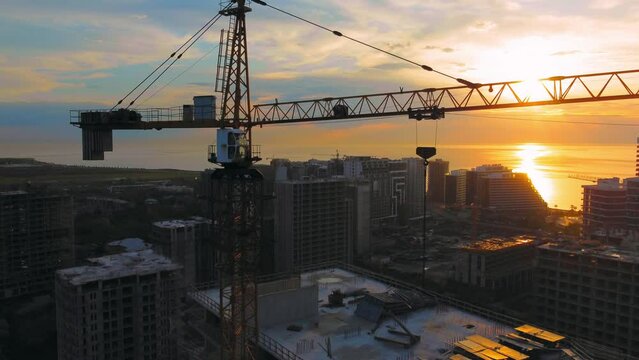 New modern apartment buildings, high-rise building with construction crane against backdrop of sunset, sun on ocean or sea. Drone view of crane construction site over city at sunset. Contractor.