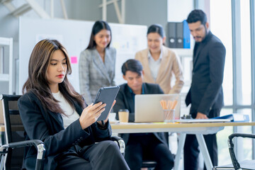 Portrait of young business woman sit on chair also hold tablet and work in office in front of group of co-worker work in the back.