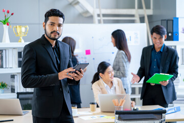 Portrait of Asian business man with beard stand with hold the tablet and look at camera also stay in front of other co-worker work in the back in office.