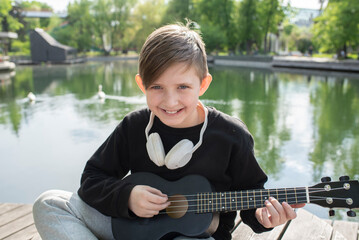 Fototapeta premium a little boy sits by the fountain, plays the black ukulele on a sunny day in the park 