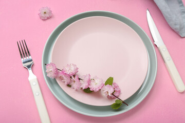 Beautiful table serving with cutlery, blooming tree branch, flowers and folded napkin on pink background