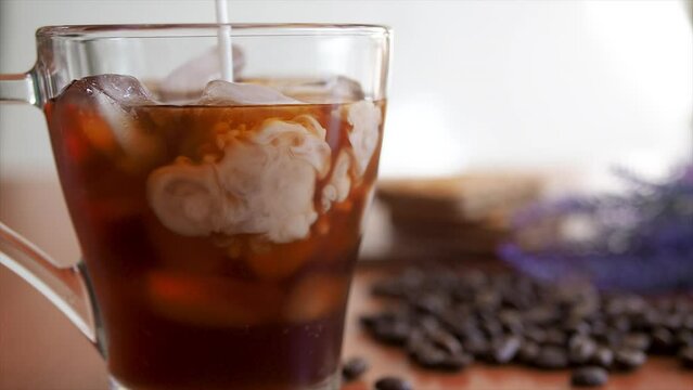 Ice coffee in a tall glass and pouring milk slowmotion shot.Cold summer drink. glass of ice and coffee beans on a table, Cookies and milk bottle on background