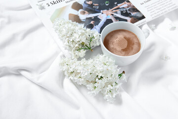 Beautiful lilac flowers with newspaper and cup of coffee on bed