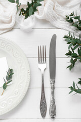 Plate with cutlery, blank card and plant branches on white wooden background