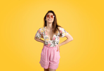 Pretty young caucasian lady wearing trendy summer outfit and colorful sunglasses, posing on yellow studio background