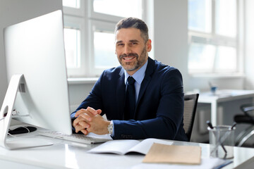 Fototapeta premium Portrait of happy middle aged businessman sitting in front of computer at workplace in office, smiling to camera