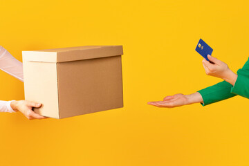 Female hands holding credit card and taking cardboard box from carrier or seller on yellow studio...