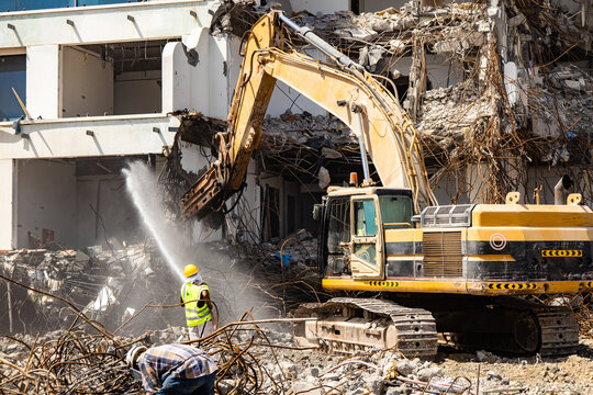 excavator equipped with a hydraulic hammer demolishes an old building while a worker sprays water to minimize dust