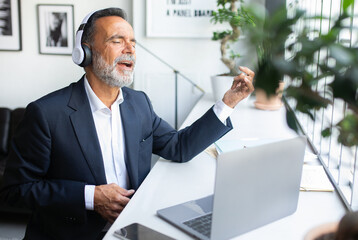 Happy old caucasian man in suit and wireless headphones resting, singing at table in office