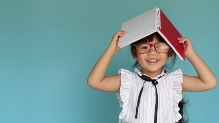 Little glasses girl with a book on her head. A cute girl laughs with book on head. Smart child with a book. 