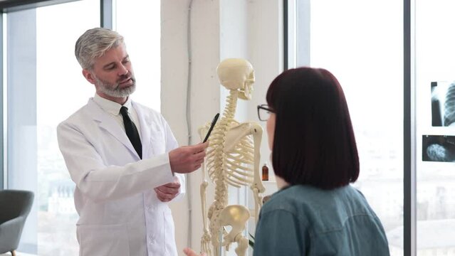 Mindful man with pen standing near anatomical model while young female sitting on exam couch in consulting room. Family doctor giving recommendations about treatment to lady with cervical brace.