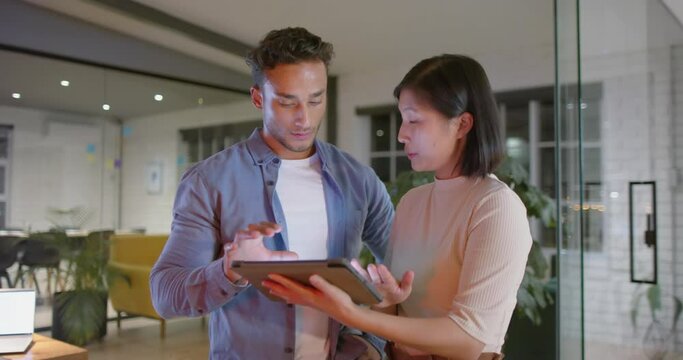 Focused diverse male and female colleague standing talking and using tablet at night in office