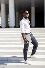 stylish African businessman in a white shirt with a laptop in his hands on the background of the building