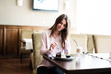 Young beautiful brunette woman sit in coffee shop cafe restaurant indoors and eat chocolate brownie dessert cake.