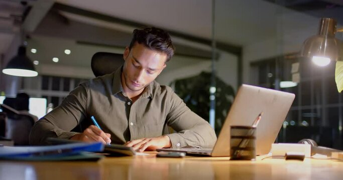 Busy biracial businessman at desk using laptop and making notes in office at night, slow motion