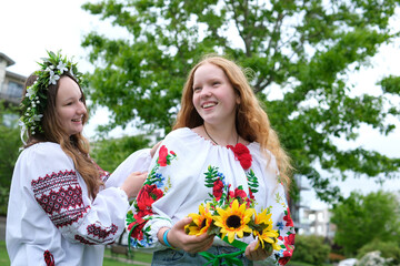 two beautiful Ukrainian girls weave wreaths embroidered shirts greenery nature sit near house...