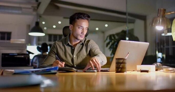 Biracial businessman at desk using laptop and smartphone in office at night, slow motion