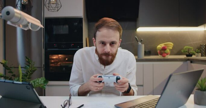 Attractive young man playing video game in moderm kitchen apartment enjoying indoor activity. Online entertainment and leisure activity concept.