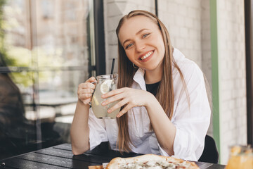 Attractive young woman with a glass of lemonade on a hot summer day. Beautiful female look dreaming and happy, she is smiling. Summer mood, 
fresh drinks. Girl sitting in cafe outside with lemonade.