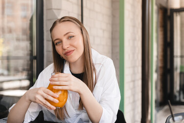 Close-up of young caucasian woman enjoying drinking orange lemonade sitting in cafe. Blonde in white shirt quenches her thirst summer. Relaxation concept. Happy laughing woman.