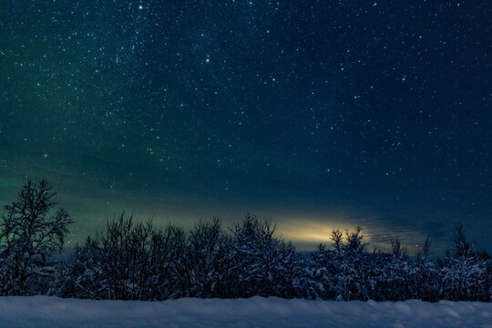 Landscape of the night sky with bright stars and green northern lights on the background of the road, trees, nature, snow in winter