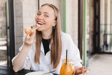 Foto auf Acrylglas Woman eating tasty pizza outdoor in street cafe. Fast food takeaway in sunny day. Blonde woman wear white shirt, orange lemonade and pizza on restaurant table. Female enjoy of delicious slice pizza. © zvkate
