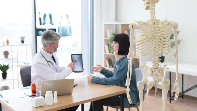 Focus on back view of teaching model located in consulting room of private practice with adults talking in background. Experts using skeleton as representation of human anatomy for patient education.