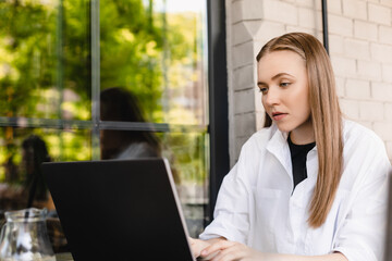 Confused young attractive blonde woman with casual hairstyle keeping hand on keyboard of laptop while looking perplexedly at screen with raised eyebrow sitting in cafe outside, serious girl working.