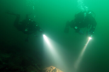 Two scuba divers pointing at an underwater stone formation with their torches