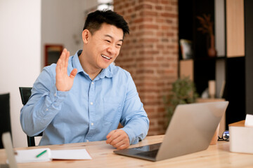 Fototapeta premium Smiling asian middle aged businessman waving hand at laptop webcamera, having online meeting at home